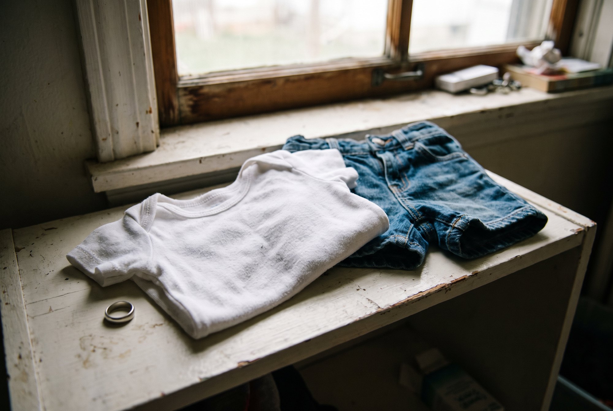 A small fitted white baby tee folded on a white shelf beside a pair of blue denim shorts and a silver ring