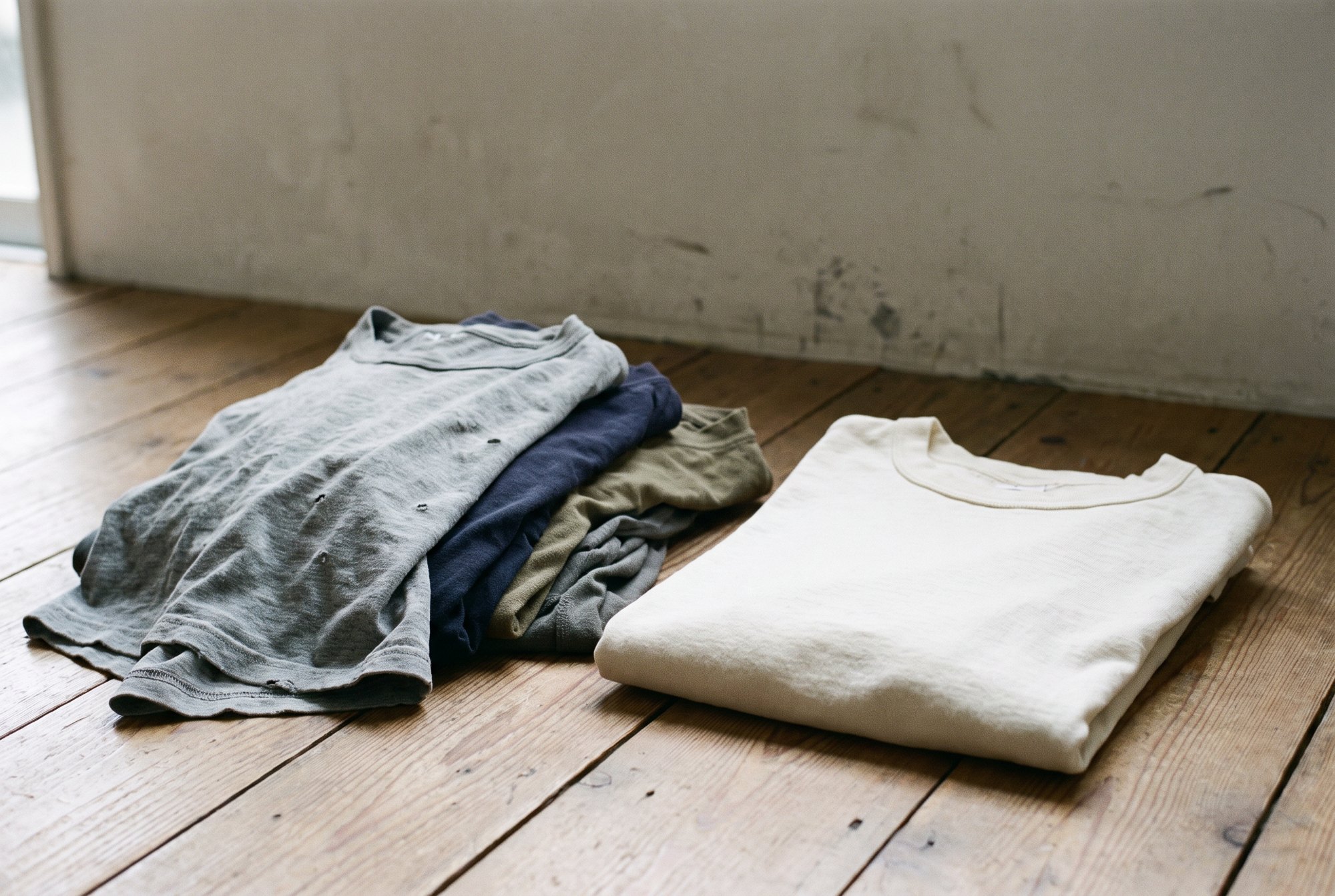A small pile of thin worn-out cheap tees on a floor next to one neatly folded thick cotton tee