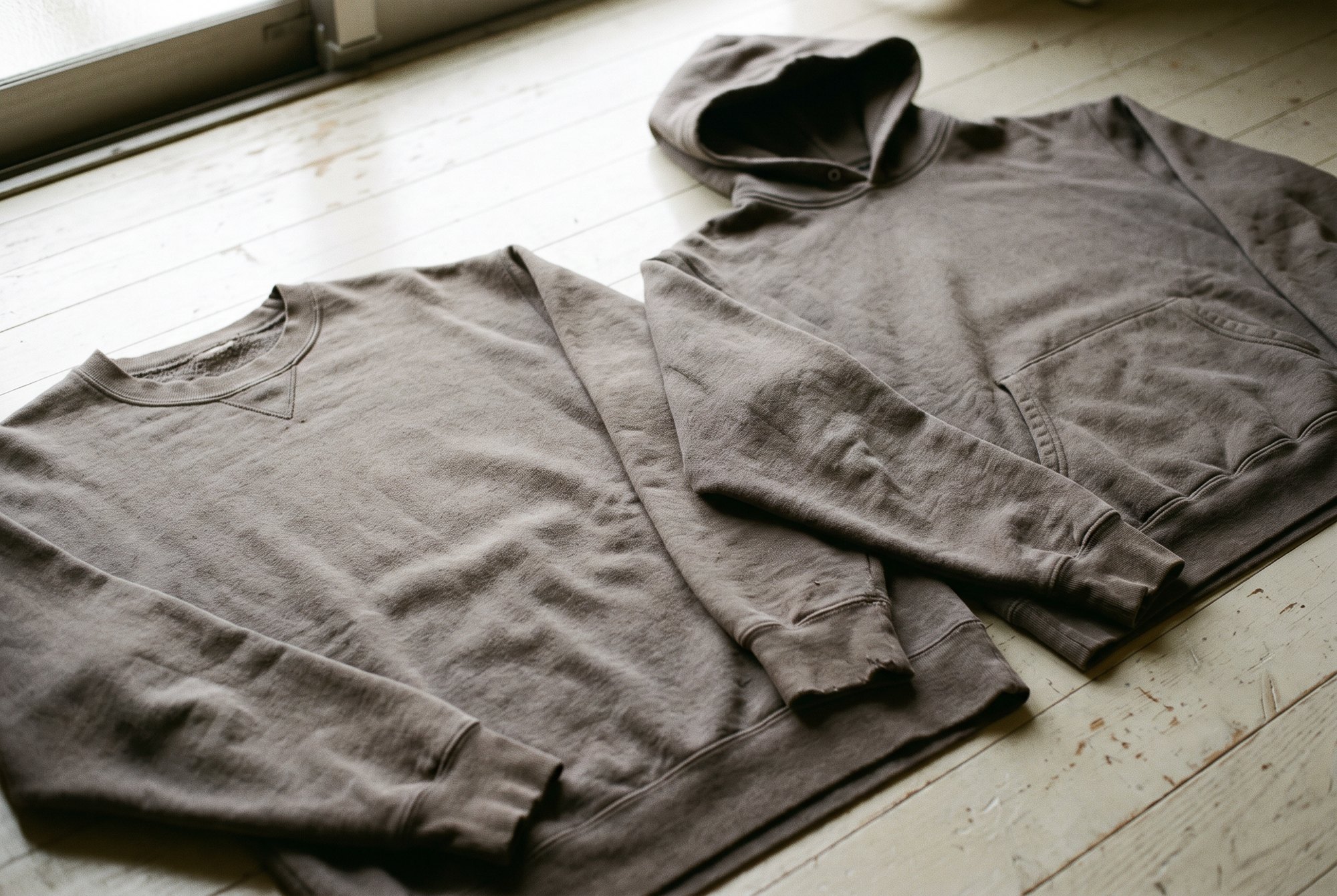 A plain grey crew sweatshirt and a plain grey hoodie laid flat next to each other on a pale floor